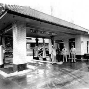 Three white male workers standing near pumps at a brick gas station under sign saying "continental oil co."