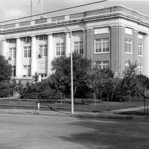 Three-story building with trees on either side of the entrance with tower in the background and car parked on its side