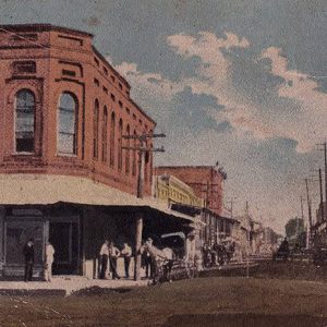 Horse drawn carriages on town street with group of people standing on street corner outside of multistory building