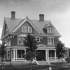 Black and white photo of three-story brick house with covered porch and brick chimneys and small trees in front