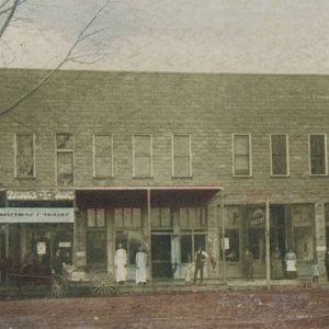 Horse drawn carriage in front of two-story brick storefront with people standing on covered sidewalk