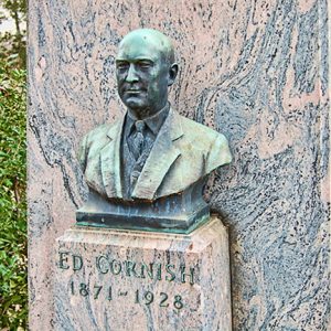 Green oxidized bust of Ed Cornish on pedestal with graveyard in backgroun