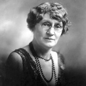 Older white woman with glasses wearing a beaded necklace and fancy dress