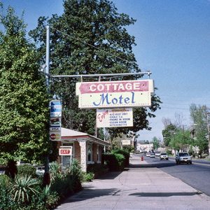 building on street with houses and cars and hanging "cottage motel" sign