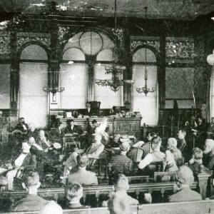 White men in crowded courtroom with arched windows and chandeliers