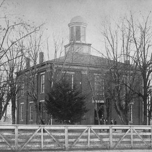 Multistory brick building with cupola and bare trees inside wooden fence