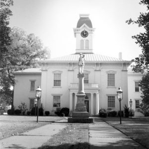 Symmetrical building with clock tower located behind stone monument with lamps on either side of it