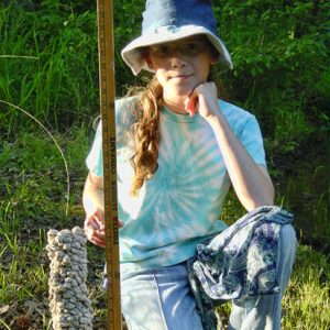 White girl in hat using a yard stick to measure height of a crayfish burrow