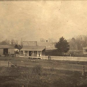 Barn and storefront with covered entrance on dirt road with fence and multistory house behind it