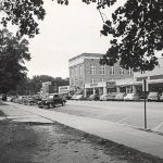 House with covered porch and parked cars across the street from row of single and multistory buildings in town