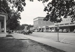 House with covered porch and parked cars across the street from row of single and multistory buildings in town