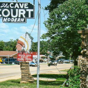 "The Cave Court modern" hanging sign and sign featuring head of Native American in feathered head dress on multilane street with storefronts in the background