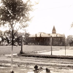 Dirt road and field with large brick building cars trees and water tower in background