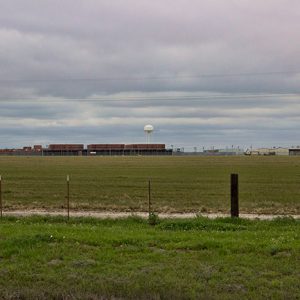 Barbed wire fence and grass field with prison buildings and water tower in the background