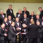 Chorus and orchestrator on stage white men in tuxedos purple vests singing with jazz hands