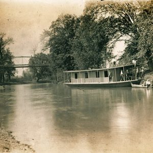 Men on houseboat on river with trees and steel arch bridge in the background