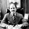 White man with mustache in suit and tie sitting at his desk with framed portrait on the wall behind him