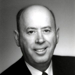 Older white man in suit standing at brick wall