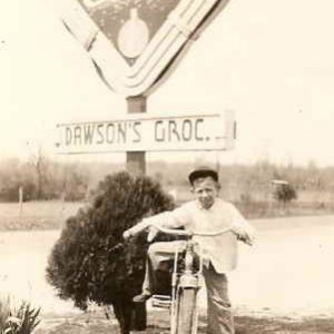 Young white boy on bicycle under a diamond shaped Coca-Cola and Dawson's Grocery sign