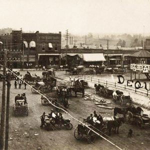 Town square crowded with horse drawn carriages and lined with brick buildings around fenced-in park with gazebo