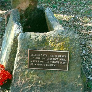 Grave with marker and flat stone blocks with plaque and flowers and a sign saying "Legend says this is grave of one of DeSoto's men. Marks on headstone may be Masonic emblem."