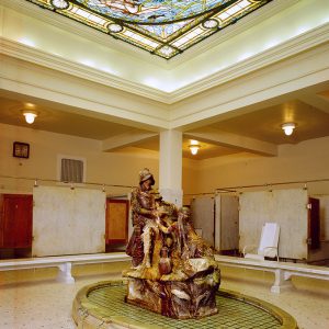 Sculpture of Native American woman pouring water for man in armor on oval base in bathing room with multiple stalls and stained glass ceiling