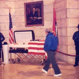 Older white man views open casket under painting with flags and police guard in State Capitol building