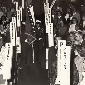 Young soldiers carrying flags in room filled with delegates sitting according to county name