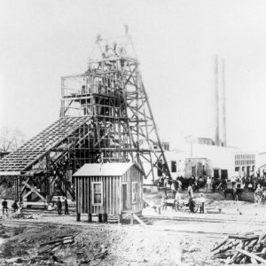 Workers and horses standing outside pit top tower and industrial buildings with smoke stacks