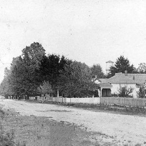 Houses with covered porches inside fence on dirt road with water tower in the background