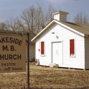 Single-story church building with cupola and wood siding with sign in the foreground