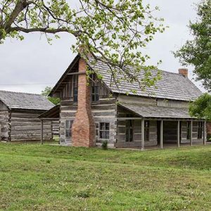 Two log cabin buildings with brick chimneys on grass with trees