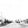 Esso gas station and storefronts on street with parked cars