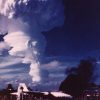 White men and airplane on landing strip with mushroom cloud in the background