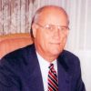 White man with glasses in suit and tie sitting and smiling in chair