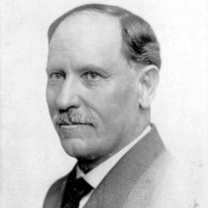 White man with mustache posing in suit and tie