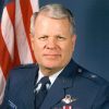 White man with gray hair in military uniform and flag behind him