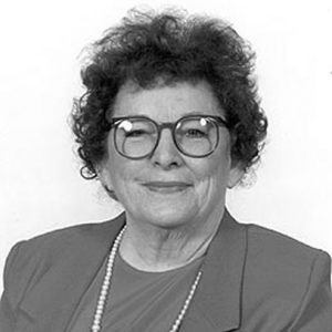 White woman smiling in glasses and suit with pearl necklace