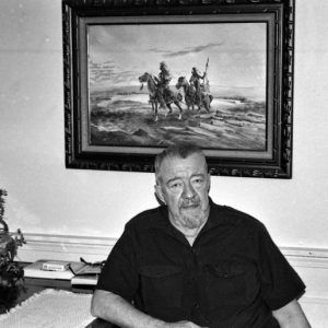 Older white man sitting at his desk with a framed painting of two Native American men on horseback hanging behind him