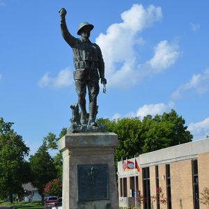 Soldier with right hand raised on pedestal with plaque in flower bed next to brick building