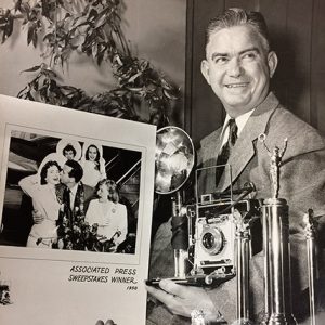 White man in suit smiling while holding camera and framed photograph with trophy in the foreground