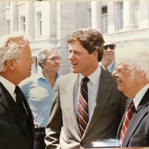 Three white men in suit and tie speaking to each other with two white men behind them outside State Capitol building