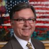 White man with glasses smiling in suit jacket and tie with flag behind him