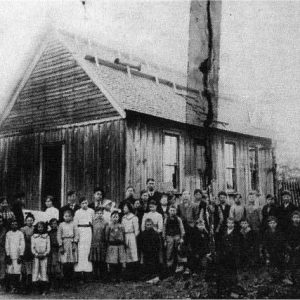 Group of white children and teachers posing for a photo outside single-story building with open gable roof