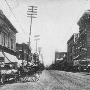 Street with trolley tracks lined with tall buildings electrical poles and lines and horse-drawn carriages