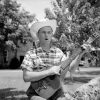 White man in cowboy hat and flannel shirt playing an acoustic guitar