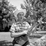 White man in cowboy hat and flannel shirt playing an acoustic guitar