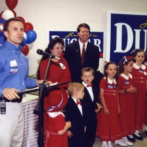 White man speaking at lectern with microphone with white man woman and children in suits and red dresses waiting beside him