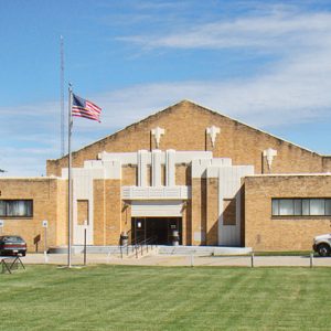 buff brick building with pointed roof and flag and lawn in front