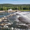 man sitting on rocks at river dam with tree covered mountain in the background